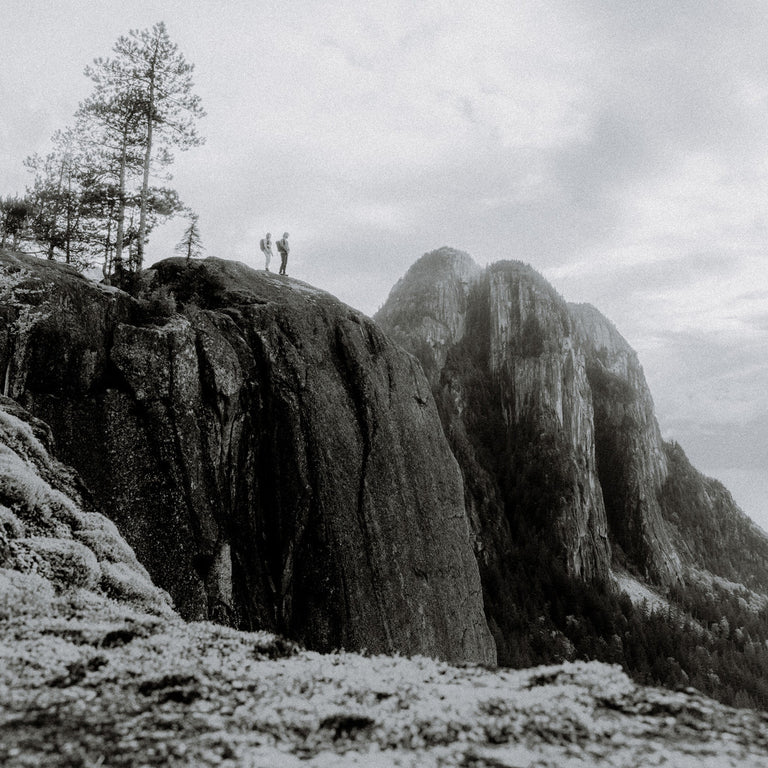 Black and white landscape photograph of towering rocky mountains under a cloudy sky, with hikers standing on a cliff edge and trees on the left.