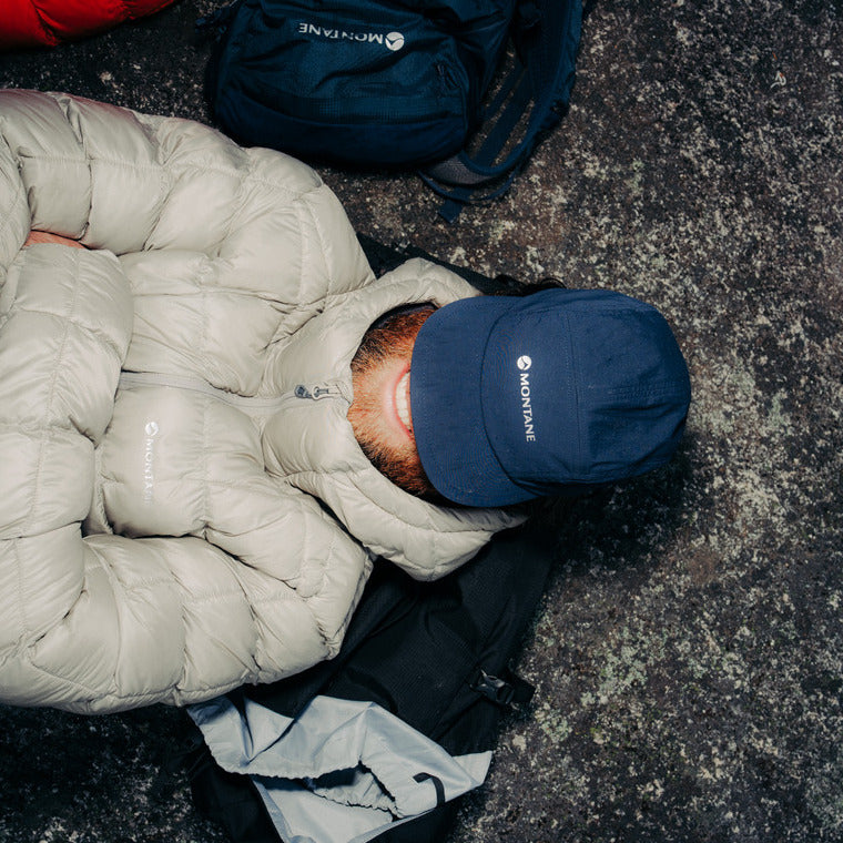 A person lying on a dark textured ground wearing a Montnae Icarus 3.0 jacket in ridge with a navy blue cap with a visible Montane logo, and a black backpack.