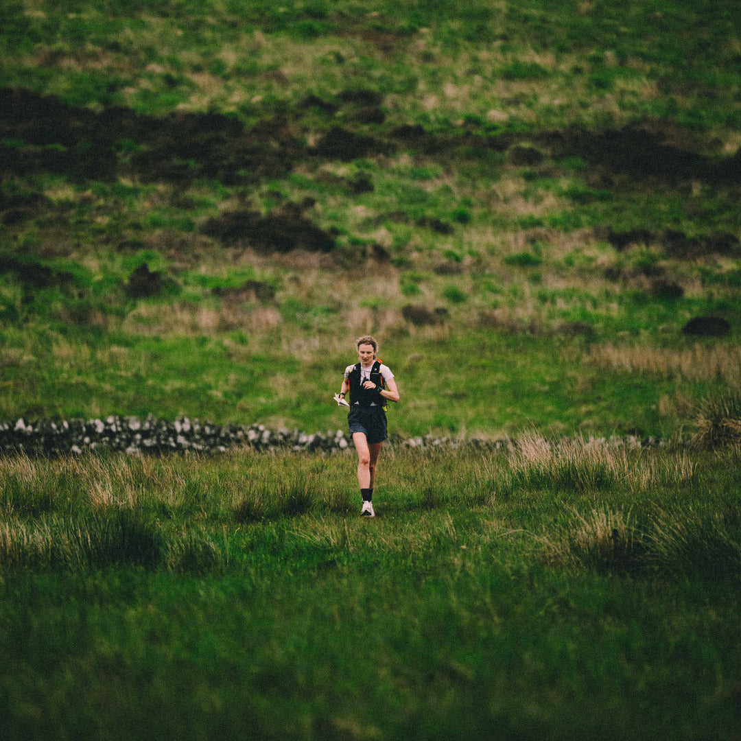 A female runner in a black athletic outfit and hydration pack running through a grassy field, with a blurred green hillside in the background.