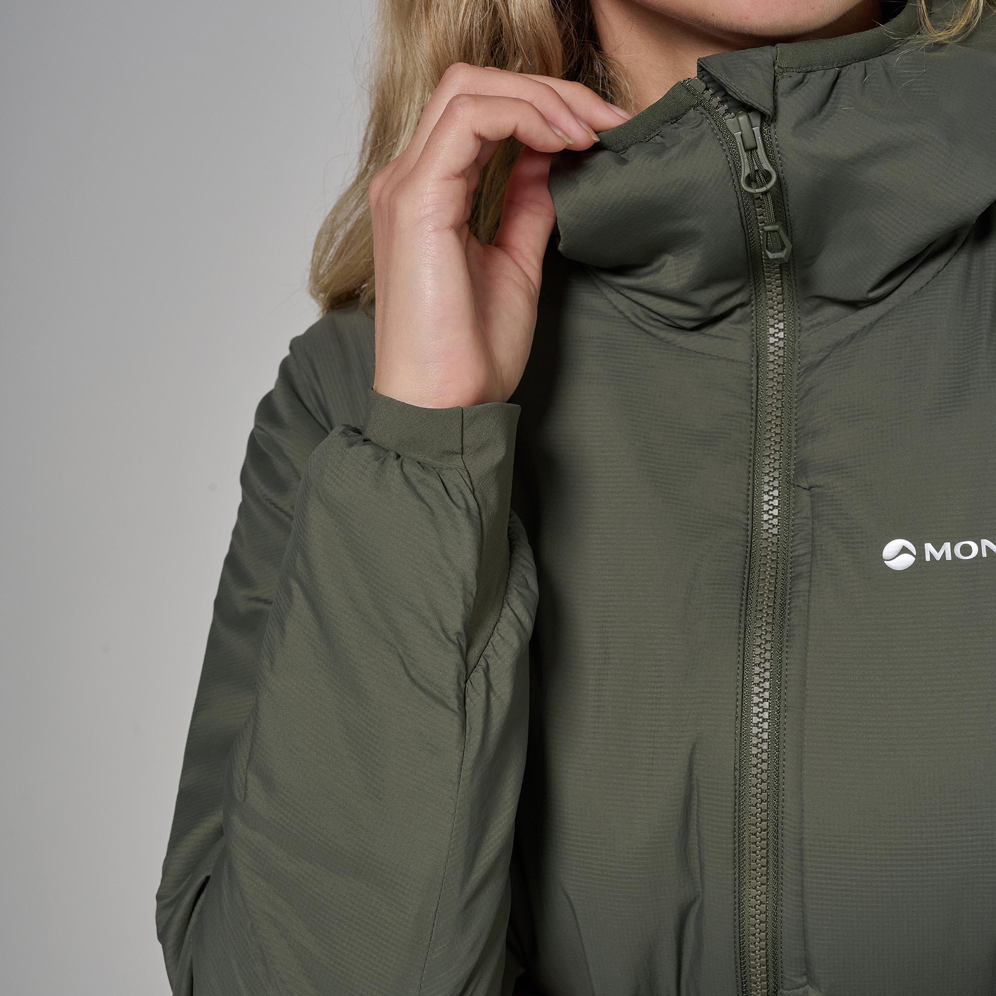 Close-up of a person wearing an olive green hooded puffer jacket, holding up the hood with one hand. The brand logo 'MON' is visible on the chest.